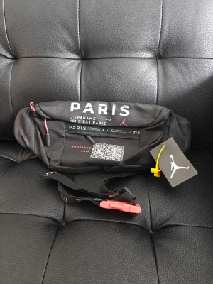 Jordan x Psg svart midjeväska  - Snygg svart midjeväska från Jordan x Paris med vita och rosa detaljer samt texten 'PARIS' och Jumpman-loggan. Väskan har justerbart band med spänne och flera fack. Perfekt för att bära det viktigaste på ett stilrent sätt.