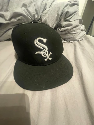Svart White Sox keps New Era 59FIFTY - Säljer en svart New Era 59FIFTY keps med Chicago White Sox-logga broderad i vitt framtill. Klassisk flat brim och officiell MLB On Field Cap. Snygg och stilren modell som passar perfekt till streetwear-looken.