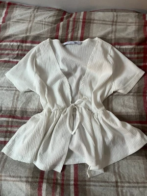 Vit topp - Säljer en vit  från lager 157 i storlek S. 💕Toppen har korta ärmar, v-ringning och ett knyt i midjan som ger en snygg siluett. Materialet är lätt och har en ribbad struktur. ☀️