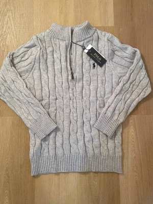 Grå stickad half zip tröja Polo Ralph Lauren - Snygg grå kabelstickad tröja från Polo Ralph Lauren med half zip och hög krage. Tröjan har långärmade ribbade muddar och klassisk broderad logga på bröstet. Perfekt för dig som gillar stilren och tidlös design.