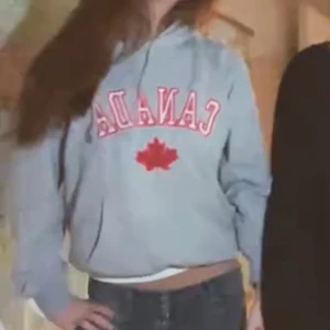 Grå hoodie med CANADA-tryck - Snygg grå hoodie med röd CANADA-text och lönnlöv på bröstet. Klassisk modell med huva och dragsko, samt magficka framtill. Perfekt för dig som gillar sportig och avslappnad stil.