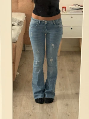 Ljusblå  bootcut jeans med slitningar - Snygga ljusblå bootcut jeans med slitna detaljer och klassisk femficksmodell. Jeansen har låg midja och är tillverkade i mjukt denimtyg som ger en avslappnad vibe och är figursydd upptill. Perfekta för dig som gillar en retroinspirerad look.
