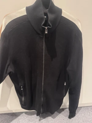 Svart ziptröja från Boss - Snygg svart ziptröja med högt krage och dragkedja framtill. Ärmarna har stickade ränder i vitt och beige/guldigt. Storlek M, nyskick!!