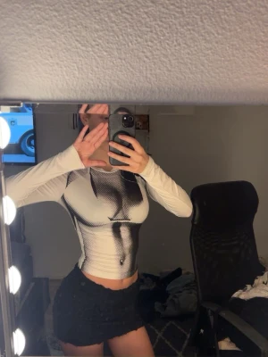 Trendig topp - Säljer en vit långärmad topp med ett unikt svart tryck framtill som ger en edgy look. Toppen är tight och croppad, perfekt att styla med höga jeans eller kjol. Materialet känns stretchigt och bekvämt. Aldrig använd