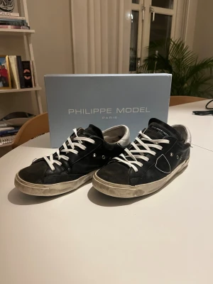  Philippe Model skor i stl 39  - Säljer ett par svarta Philippe Model sneakers med vita snören och grå detaljer vid hälen. Skorna har en klassisk låg profil, rund tå och platt sula. Tillverkade i skinn med snygga sömmar och metallnitar på sidan. Perfekta för dig som gillar stilrena och trendiga sneakers. Haft de i ett tag men använt de sparsamt. De är nu uppfräschade med nya snören och tvättade. Modellen är PM/78 EDT. 3500kr nya