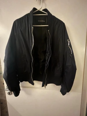 Svart bomberjacka från Rut&Circle - Stilren svart bomberjacka från Rut&Circle med klassisk ribbad krage, muddar och dragkedja framtill. Jackan har fickor på sidorna och en extra ficka med dragkedja på ärmen. Perfekt för dig som vill ha en enkel men snygg look.