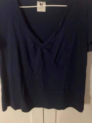 Mörkblå v-ringad topp från Nanso - En enkel och stilren mörkblå topp från Nanso med v-ringning och korta ärmar. Toppen har en lätt rynkning vid ringningen som ger en snygg detalj. Perfekt att matcha med jeans eller kjol för en clean look.