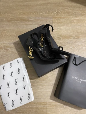 Svarta med logoklack finns olika storlekar  - Säljer ett par svarta pumps i lack med öppen tå och ikonisk guld YSL-logga som klack. Skorna har en elegant, spetsig form och tunna remmar över foten. Perfekta för dig som vill sticka ut med lyxig design.