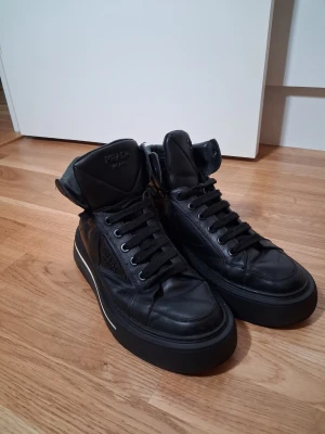 Svarta boots från Prada i skinn - Snygga svarta boots från Prada med högre skaft och klassisk snörning. Tillverkade i skinn med tydlig Prada-logga på sidan och bak på sulan. Skorna har en platt sula och en modern, chunky design som passar till många olika stilar.