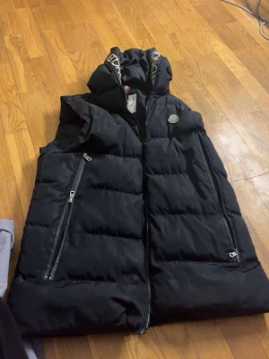 Svart dunväst från Moncler - Svart dunväst från Moncler med huva och dragkedjor på sidorna. Västen har Moncler-logga på bröstet och tryckt text på huvan. Perfekt för lager-på-lager och håller dig varm under kyliga dagar.