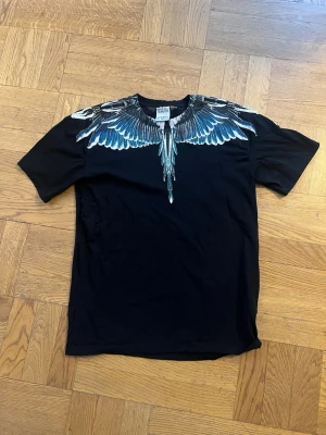 Svart t-shirt med vingeprint från Marcelo Burlon - Svart t-shirt från Marcelo Burlon med ikoniskt vingeprint i blått, vitt och grått runt halsen både fram och bak. Klassisk passform och rund halsringning. Perfekt för dig som vill sticka ut med en unik detalj.