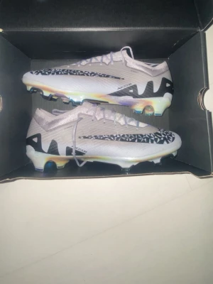 Nike Zoom Mercurial fotbollsskor grå - Nike Zoom Mercurial fotbollsskor i ljusgrå färg med svarta detaljer och stor Swoosh-logga på sidan. Skorna har snörning, platt sula med dobbar och en cool iriserande undersida med rosa inslag. Tillverkade i syntetmaterial för bästa prestanda på planen. Pris kan diskuteras 