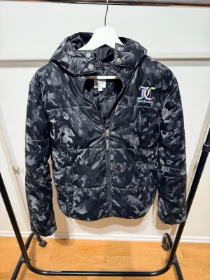 Svart camo pufferjacka Juicy Couture - Svart pufferjacka från Juicy Couture med camouflagemönster och huva. Jackan har dragkedja framtill, två stora fickor och broderad logga på bröstet. Perfekt för kalla dagar när du vill ha en cool streetstil.