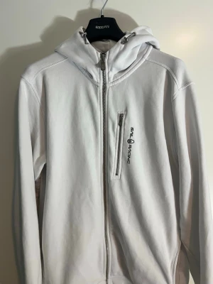Bowman Vit hoodie från Sail Racing - Nästan som ny. Vit hoodie med dragkedja från Sail Racing. Har en bröstficka med dragkedja och logga broderad i svart på vänster sida. Utrustad med huva och detaljerad sömnad på axlar och huva. Tillverkad i mjukt bomullsmaterial för en clean och sportig look. Nypris 1500 kr