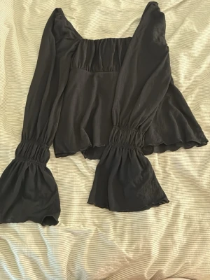 Svart blus med vida ärmar - Trendig svart blus med vida, långa ärmar och smockdetaljer vid ärmslut och axlar. Blusen har en lös passform och är tillverkad i ett lätt tyg som ger ett snyggt fall. Perfekt för dig som gillar en stilren men ändå unik look.