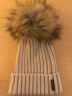 Beige stickad mössa med pälsboll Hollies - Snygg beige ribbstickad mössa från Hollies med stor fluffig pälsboll i bruna och svarta toner. Mössan har uppvikt kant och en liten svart logotypetikett. Perfekt accessoar för kalla dagar och trendig vinterstil.