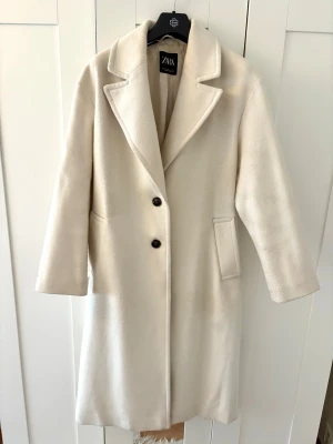krämvit kappa från Zara med knappar - Stilren beige kappa från Zara med klassisk krage och två svarta knappar framtill. Kappan har raka ärmar, två fickor och en rak passform som ger en clean look. Perfekt för dig som gillar minimalistisk stil och vill ha en tidlös jacka i garderoben.