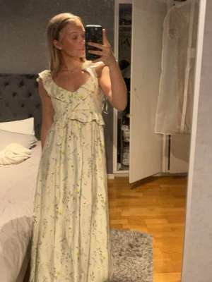 Blommig maxiklänning med volang - Söt maxiklänning i ljus färg med blommigt mönster och volangdetaljer vid axlar och byst. Klänningen har ärmlös design och v-ringning, perfekt för varma dagar och somriga vibes.