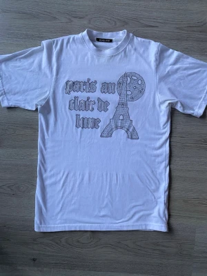 Vit t-shirt med Paris-tryck från BLINGLUCD - Vit t-shirt i bomull med tryck framtill som föreställer Eiffeltornet, måne och texten 'paris au clair de lune' i coolt typsnitt. Klassisk passform med rund hals och korta ärmar. Perfekt för dig som gillar franska vibbar och streetstyle. Alla pärlor kvar
