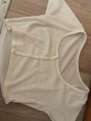 Beige ribbad kortärmad topp - Kortärmad topp i beige ribbat material med rund halsringning och knappar framtill. Croppad modell som passar perfekt till höga jeans eller kjol. Enkel och stilren design.