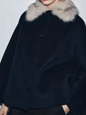 Mörkblå kappa med fuskpäls-krage - Stilren mörkblå kappa med bred, ljus fuskpäls-krage och knäppning framtill. Kappan har en loose passform och är perfekt för kyliga dagar. Enkel design med diskreta fickor och klassisk look.