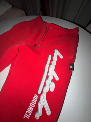 Röda mjukisbyxor från Hoodrich - Snygga röda mjukisbyxor från Hoodrich med vit broderad logga längs benet och snörning i midjan. Byxorna har en relaxed fit och är tillverkade i mjukt bomullsmaterial. Perfekta för en chill och streetwear-inspirerad look.