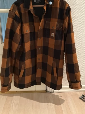 Rutig overshirt från Carhartt - Snygg overshirt från Carhartt i svart och brunrutig design. Klassisk skjortkrage, tryckknappar framtill och två bröstfickor, varav en med Carhartt-logga. Tillverkad i kraftig bomull, perfekt för lager på lager-stil.