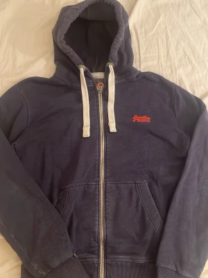 Mörkblå hoodie från Superdry - Mörkblå hoodie med dragkedja från Superdry Orange Label. Har vit dragsko i huvan, två fickor fram och broderad logga i rött på bröstet. Mjuk insida och ribbade muddar. Perfekt för en chill och avslappnad stil.