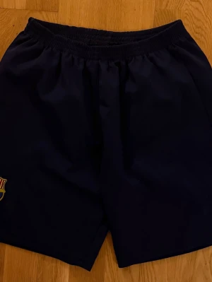 Blå FC Barcelona shorts - Mörkblå shorts med FC Barcelona-logga på ena benet. Klassisk sportig design med resår i midjan, perfekta för fotboll eller träning.