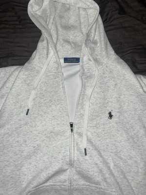 Ljusgrå hoodie från Polo Ralph Lauren - Snygg ljusgrå hoodie från Polo Ralph Lauren med dragkedja framtill och klassisk huva med dragsko. Broderad marinblå logga på bröstet och känguruficka. Tillverkad i mjukt bomullsmaterial som är perfekt för chill dagar.