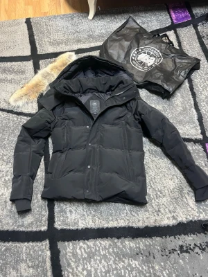 Svart dunjacka från Canada Goose - Snygg svart dunjacka från Canada Goose med avtagbar huva och pälskant. Jackan har quiltad design, dragkedja och knappar framtill samt långa ärmar med muddar. Perfekt för kalla vinterdagar och riktigt stilren look.