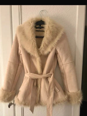 Beige pälsjacka - Snygg beige pälsjacka med fuskpäls runt krage, ärmslut och nederkant. Jackan har knappar framtill och ett matchande bälte i midjan. Väldigt elegant. Enbart använd en gång❤️