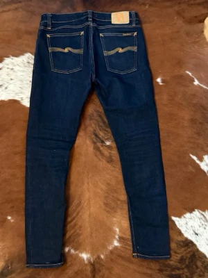 Mörkblå jeans från nudie - Mörkblå jeans från Nudie med klassiska kontrastsömmar. Jeansen har fem fickor och Nudie-logga på bakfickorna. Perfekta för dig som gillar en stilren och enkel look. Passar w31