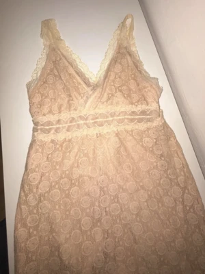 Beige spetsnattlinne med v-ringning - Sött nattlinne i beige spets med v-ringning och smala axelband. Spetsen har ett cirkelmönster och nattlinnet är dekorerat med spetskant längs urringningen och midjan. Perfekt för en mysig kväll hemma.