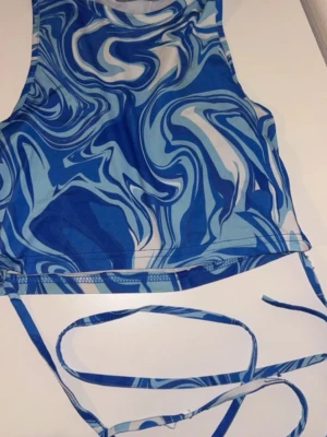 Blå swirl topp från Zechka - Säljer en blå och vit swirlmönstrad croppad topp från Zechka. Toppen har smala axelband och långa knytband i midjan som kan stylas på olika sätt. Materialet är mjukt och stretchigt, troligen polyester. Perfekt för en trendig look!