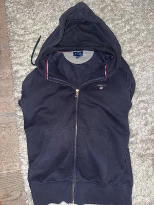 Mörkblå hoodie från Gant med dragkedja, bra skick, original pris 1300 - Snygg mörkblå hoodie från Gant med dragkedja framtill och två stora fickor. Klassisk huva med dragsko och diskret Gant-logga broderad på bröstet. Mjuk bomullsblandning och ribbade muddar. Perfekt för en avslappnad och sportig stil.