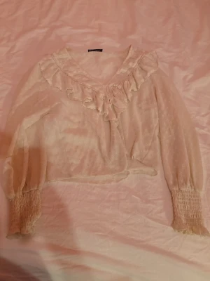 Beige volangblus med prickigt mönster - Söt beige blus från Bershka med volangdetaljer längs v-ringningen och diskret prickigt mönster. Blusen har långa, transparenta ärmar med smock vid ärmsluten och är tillverkad i ett lätt, luftigt tyg. Perfekt för en trendig och romantisk look.