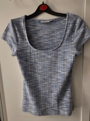 Blåmelerad ribbad topp från Kappahl - Kortärmad topp i blåmelerat ribbat tyg med rund, djup urringning. Snygg och enkel att matcha till olika outfits. Materialet känns mjukt och stretchigt, perfekt för en avslappnad look.