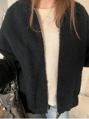 Svart teddyjacka med dragkedja - Mysig svart teddyjacka med dragkedja framtill. Jackan har en enkel, stilren design och är perfekt för att hålla värmen under kyliga dagar. Passformen är något loose och materialet är fluffigt och mjukt, vilket ger en avslappnad vibe.