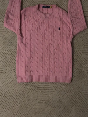Rosa kabelstickad tröja Polo Ralph Lauren - Kabelstickad rosa tröja från Polo Ralph Lauren med rund halsringning och broderad logga på bröstet. Tröjan har ribbade muddar vid ärmslut och nederkant. Perfekt för dig som gillar klassisk och stilren look.