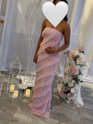 Ljusrosa glittrig långklänning - Superfin ljusrosa långklänning med glittriga paljetter och pärlor. Klänningen är axelbandslös och har en figurnära passform med volangdetaljer i lager. Perfekt för bal eller festliga tillfällen.