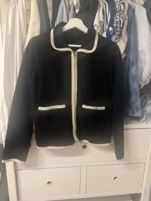 Svart kavaj med detaljer - Stilren svart kavaj med raka linjer och dekorativa detaljer längs kanter, fickor och ärmslut. Kavajen har två fickor framtill och en klassisk krage. Perfekt för en trendig och elegant look.