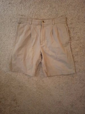 Beige bomullsshorts med veck - Stilrena beige shorts i bomull med klassiska pressveck framtill, bälteshällor och knappstängning. Perfekta för sommaren och lätta att matcha med olika plagg.