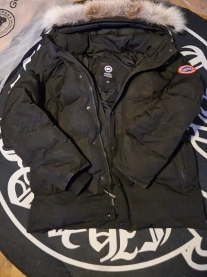 Svart dunjacka från Canada Goose XL - Svart dunjacka från Canada Goose med pälskantad huva och klassisk logga på ärmen. Jackan har dragkedja framtill och är perfekt för kalla vinterdagar. Snygg och tidlös design som håller dig varm hela säsongen. Säljes pga för stor 