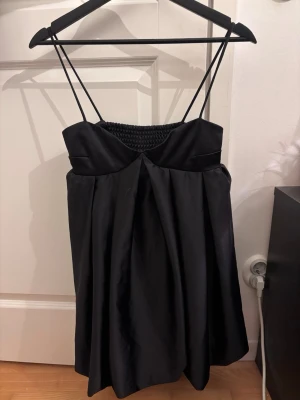 Svart kort klänning med tunna axelband - Stilren svart kort klänning med tunna axelband och smockad rygg. Klänningen har en lätt utsvängd form och är tillverkad i ett glansigt material som ger en elegant känsla. Perfekt för dig som vill ha en enkel men snygg look.