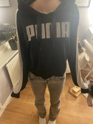 Svart hoodie från Puma med tryck - Snygg svart hoodie från Puma med stort vitt logotryck på bröstet och vita detaljer längs ärmarna. Klassisk huva med dragsko och avslappnad passform. Perfekt för en chill och sportig stil.