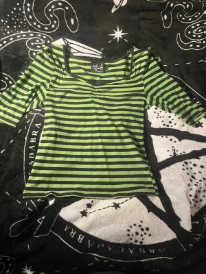 Grön och svart randig topp Hell Bunny - Snygg topp från Hell Bunny i grönt och svart med horisontella ränder. Modellen har trekvartsärm, v-ringning och en liten rynkning vid bysten. Perfekt för dig som gillar alternativ stil och vill sticka ut lite extra. En sömm har släppt i nedre kanten men inget som syns eller inte går att fixa.
