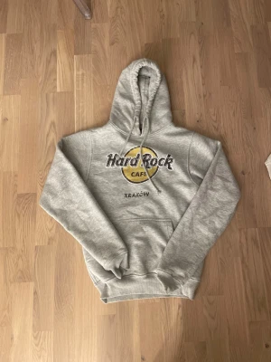 Grå Hard Rock Cafe hoodie Kraków - Grå hoodie från Hard Rock Cafe med stor logga och texten Kraków på bröstet. Klassisk känguruficka framtill och justerbar huva med snörning. Tillverkad i mjuk bomull och polyester för skön känsla. Perfekt för dig som gillar ikoniska tryck och avslappnad stil.