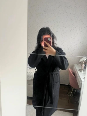 Svart kappa  - Stilren svart kappa med bälte/band  i midjan och klassisk krage. Kappan har lång ärm och rak passform.