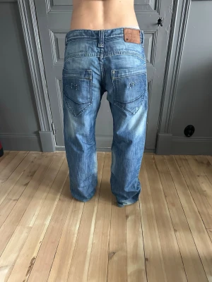 Blå loose fit jeans med slitningar - Säljer ett par blå jeans från bragf med klassisk femficksdesign och ljusa slitningar. Jeansen har en loose passform och raka ben, samt en brun läderpatch bak i midjan. Perfekta för en avslappnad stil. Midjemått: 41 cm, Innerbenslängd: 79 cm🤗